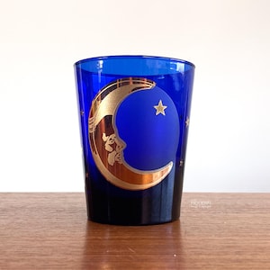 May include: A cobalt blue glass with a gold crescent moon and star design. The moon has a face etched into it. The glass is cylindrical and sits on a wooden surface. The glass is a decorative item.