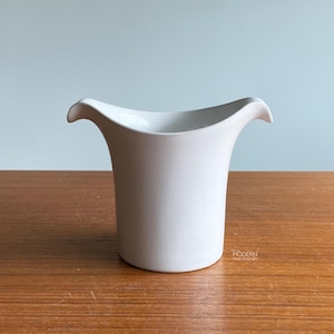 May include: A white ceramic creamer or small pitcher with a smooth, matte finish. The vessel features a wide, flared rim with two distinct, symmetrical pouring spouts on opposite sides, creating a curved, wing-like silhouette at the top.