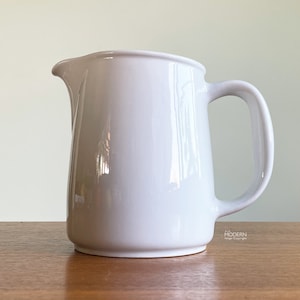 May include: A white ceramic pitcher with a curved handle and a spout. The pitcher has a smooth, glossy finish and sits on a wooden surface. The design is simple and modern, suitable for serving beverages.