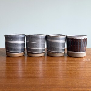 May include: Four cylindrical ceramic cups arranged in a row. The first three cups feature horizontal gray and white marbled patterns with unglazed tan bases. The fourth cup has a dark brown vertical ribbed texture with a smooth white rim and base.