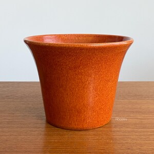 May include: A flared ceramic planter with a vibrant orange speckled glaze and a smooth finish. The exterior features a subtle, small white stamp near the base that reads ASA MODERN, indicating the brand of this round, open-top vessel.
