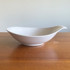 May include: A white ceramic serving bowl with a unique, curved design. The bowl has a wide, oval shape with a raised, flared rim on one side. The bowl is set on a wooden surface.