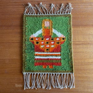 May include: A rectangular woven wall hanging with a green background and a central figure in orange and red. The figure has a white top and is surrounded by a brown border. The bottom edge has white fringe.