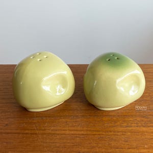 May include: A pair of ceramic salt and pepper shakers. One shaker is a pale yellow, the other is a pale yellow with a green top. Each shaker has three holes on top for dispensing. The shakers have a rounded, organic shape.