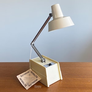 May include: Cream-colored Mobilite desk lamp with adjustable arm and shade. The lamp has a rectangular base with a switch and a tag that reads "Mobilite". The lamp is on a wooden surface.