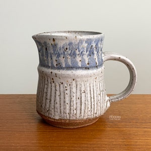 May include: A handmade ceramic pitcher with a light blue and white speckled glaze. The pitcher has a handle and a spout for pouring. The lower portion features vertical textured lines, and the base is a natural brown color.