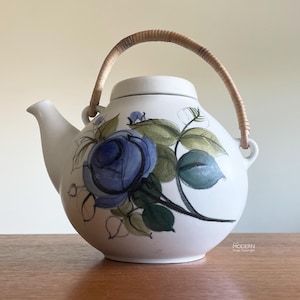 May include: White ceramic teapot with a blue rose floral design and a woven handle. The teapot features a rounded body, a spout, and a lid. The floral design includes blue roses and green leaves. The handle is made of woven light brown material.