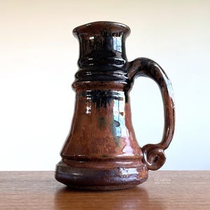 May include: A handcrafted ceramic mug with a dark brown and black glaze. The mug features a large handle and a flared top. The base is a darker shade of brown. The mug is sitting on a wooden surface.