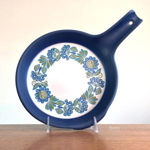 May include: A round, blue ceramic serving dish with a handle. The center features a white background with a floral design in blue and green. The handle has a hole for hanging.