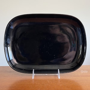 May include: A rectangular black serving tray with rounded corners. The tray has a glossy finish and is displayed on a clear acrylic stand. The tray is empty and reflects the surrounding environment. The tray is a vintage piece.