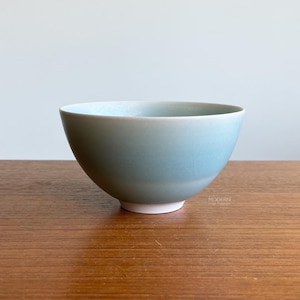 May include: A light blue ceramic bowl with a white base. The bowl has a smooth, rounded shape and a glossy finish. The bowl is sitting on a wooden surface.