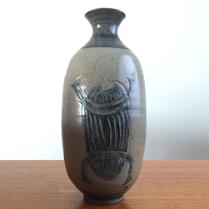 May include: A tall, cylindrical ceramic vase with a narrow neck and flared rim. The body features a speckled beige glaze with a dark, incised scarab beetle motif. The base and neck are accented with dark gray tones, and the text "AIA MODERN" is visible.