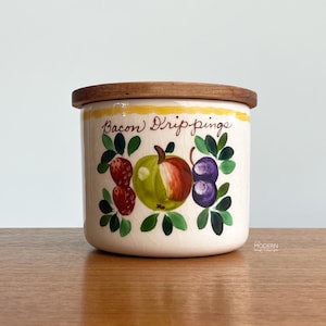 May include: A vintage ceramic bacon drippings jar with a wooden lid. The jar is decorated with hand-painted fruit illustrations, including strawberries, an apple, and plums. The words "Bacon Drippings" are written in script.