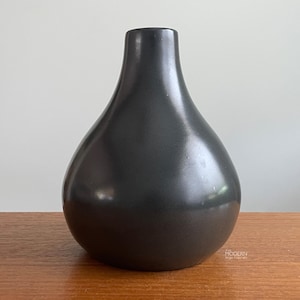 May include: A dark charcoal gray ceramic vase with a smooth, matte finish and a rounded, bulbous body that tapers into a narrow neck. The surface has a subtle sheen, and the small text "MODERN" is printed in white near the base of the object.