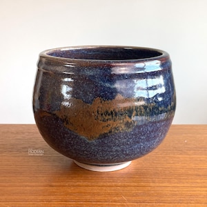 May include: A handcrafted ceramic bowl with a rounded shape and a dark blue glaze. The bowl features a band of brown and gold near the center, with a white base. The bowl sits on a wooden surface.