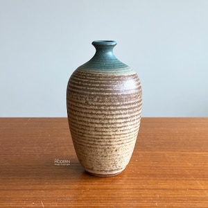 May include: A ceramic vase with a textured, ribbed design. The vase has a rounded body and a narrow neck. The top is teal, transitioning to a brown and tan color. The vase is displayed on a wooden surface.