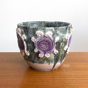 May include: A decorative ceramic planter with a textured, mottled finish in shades of green, blue, and purple. The planter features raised, ornate purple and white floral designs around the exterior. The base has a pink, vertical striped pattern.