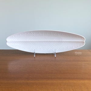 May include: A long, white, oval-shaped ceramic serving platter with a central ridge and a textured pattern of diagonal lines and small raised dots resembling a leaf. The platter is displayed on a clear acrylic stand for elevated viewing.