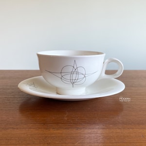 May include: A white ceramic teacup and saucer set featuring a dark brown, thin, abstract line drawing of a symmetrical, star-like geometric pattern on the side of the cup and a matching smaller design on the edge of the saucer.