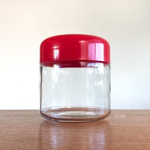 May include: A clear glass jar with a rounded base and a bright red lid. The jar is empty and sits on a wooden surface. The lid is a solid, glossy red color. The jar is a simple, cylindrical shape.