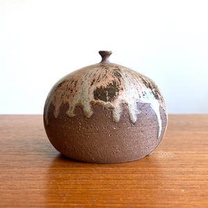 May include: A handcrafted, round ceramic vase with a dark brown base and a lighter, textured top. The top features a dripped glaze effect in shades of beige and white, adding a unique, artistic touch to the decorative piece.
