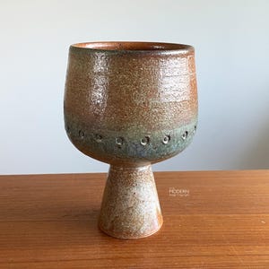May include: A handcrafted ceramic goblet with a wide, textured bowl and a pedestal base. The goblet features a brown and tan color scheme with a band of small holes around the middle. The goblet is displayed on a wooden surface.
