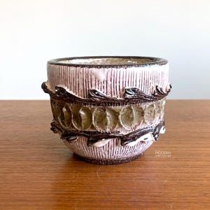 May include: A small, ceramic vessel with a textured, vertical-lined exterior in pale pink. A band of olive green, circular designs is framed by dark brown, leaf-like accents. The rim and base are also dark brown.