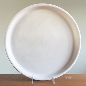 May include: A large, round, off-white ceramic serving platter. The platter has a slightly raised edge and sits on a clear acrylic stand. The surface of the platter is smooth and appears to be unglazed.