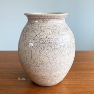 May include: A beige ceramic vase with a crackle glaze finish. The vase has a rounded body and a slightly flared rim. The surface is covered in a network of fine, dark lines, creating a unique texture. The vase is a decorative item.