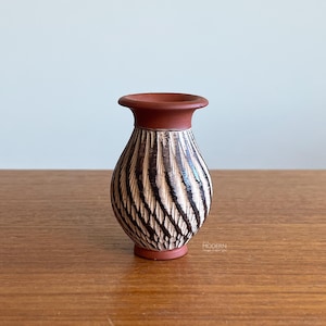 May include: A small, decorative vase with a terracotta-colored rim and base. The body of the vase features a textured pattern of alternating dark brown and beige vertical lines. The vase is displayed on a wooden surface.