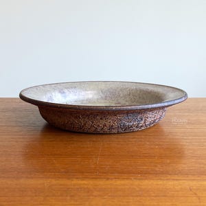 May include: A shallow ceramic bowl featuring a textured, dark brown exterior with a cross-hatched pattern and a smooth, speckled light beige interior glaze. The rim is dark brown, and the text "MID MOD MODERN" is visible on the side.