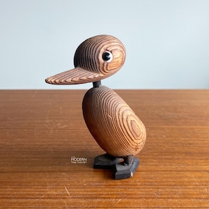 May include: Wooden duck figurine with a layered design. The duck has a brown body, head, and beak, with a black base and a single white eye. The figurine is a decorative item.