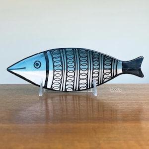 May include: A decorative glass fish-shaped dish with a blue head and tail, and a black and white patterned body. The dish is displayed on a clear stand, showcasing its design. The fish-shaped dish is a unique home decor item.