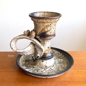 May include: A handcrafted ceramic candlestick holder with a unique design. The holder features a speckled beige and brown finish, a curved handle, and a circular base with a leaf pattern. The rim is dark brown.