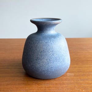 May include: A small, dark blue ceramic vase with a textured surface. The vase has a rounded body and a narrow neck with a flared rim. The color gradient transitions from a darker base to a lighter blue at the top. The vase is displayed on a wooden surface.