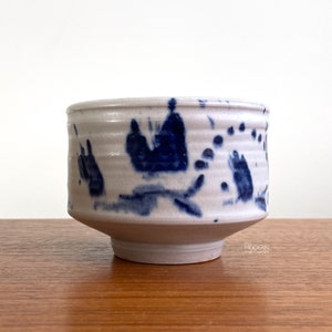 May include: A handmade ceramic bowl with a white base and blue abstract designs. The bowl has a slightly flared shape and a footed base. The blue designs appear to be brushstrokes or splatters, adding a unique touch to the piece.