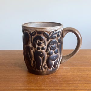 May include: A brown ceramic mug featuring a relief design of stylized figures and foliage. The mug has a smooth, curved handle and a speckled, earthy glaze. The surface displays a repeating pattern of human-like forms integrated with leaves.