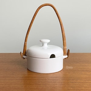 May include: A white ceramic sugar bowl with a matching lid and a tall, arched handle wrapped in light brown rattan. The bowl features a small notch on the rim for a spoon and a smooth, glossy finish. The design is simple, round, and functional.