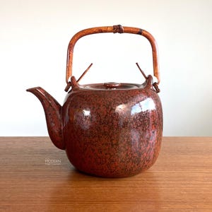 May include: A vintage-style teapot with a speckled reddish-brown glaze. It features a curved spout, a lid, and a bamboo handle. The teapot is sitting on a wooden surface.