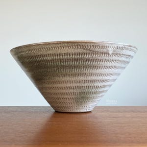 May include: A large, textured ceramic bowl with a wide opening and a conical shape. The bowl features horizontal, ridged detailing in shades of brown and cream. The bowl is displayed on a wooden surface.