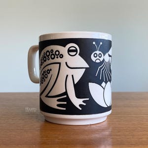 May include: A white ceramic mug featuring a bold black graphic print of a stylized frog with circular patterns on its back, positioned next to a small insect and floral shapes. The mug has a simple handle and a smooth, cylindrical body design.