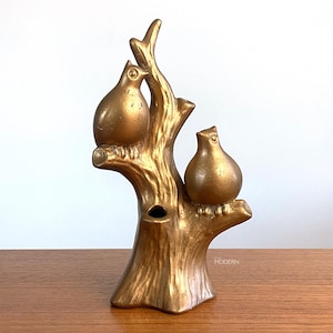 May include: A gold-colored ceramic sculpture featuring two rounded birds perched on a textured tree branch. The tree trunk has a hollow opening at the top, and the small text "AA MODERN" is visible near the base of the right side of the tree.