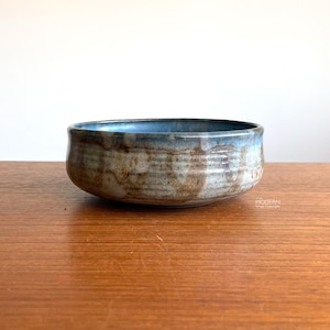 May include: A handmade ceramic bowl with a rustic, textured appearance. The bowl features a wide, shallow design with a blue-glazed interior and a mottled, earthy exterior. The bowl is set on a wooden surface.