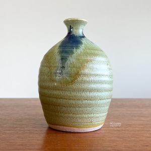 May include: A ceramic vase with a rounded body and a narrow neck, featuring a textured, ribbed surface. The glaze is primarily a mottled olive green with a prominent vertical streak of deep blue running down from the rim. The base is unglazed.