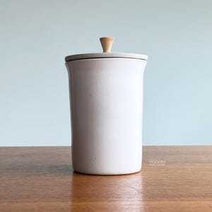 May include: A white ceramic canister with a gray lid and a wooden knob. The cylindrical container sits on a wooden surface against a light blue background. The design is minimalist and modern.