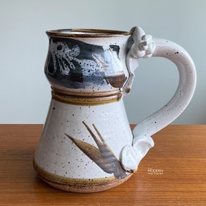 May include: A speckled ceramic mug with a flared base and a wide, curved handle featuring decorative molded floral accents. The mug has a white glaze with dark brown and black abstract brushwork, brown rim detailing, and a natural clay base.