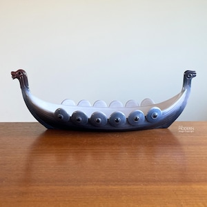 May include: A ceramic Viking ship-shaped decorative bowl. The vessel is primarily white with gradient shading to dark blue. It features a dragon head at each end and a row of shields along the sides. The bowl is displayed on a wooden surface.