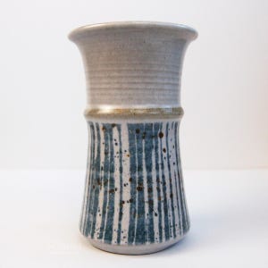 May include: A ceramic vase with a flared rim and a cylindrical body. The upper section is a smooth, light gray, while the lower section features vertical blue stripes and dark brown speckles over a light gray base, separated by a thin tan band.