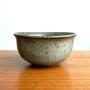 May include: A handcrafted ceramic bowl with a speckled, muted green glaze. The bowl has a rounded shape with a slightly flared rim. The interior and rim have a darker, brownish hue. The bowl is sitting on a wooden surface.