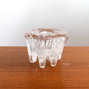 May include: A clear glass candle holder with a textured, organic design. The candle holder has a circular top with a central opening for a candle and is supported by three short legs. The glass has a rippled, textured surface.